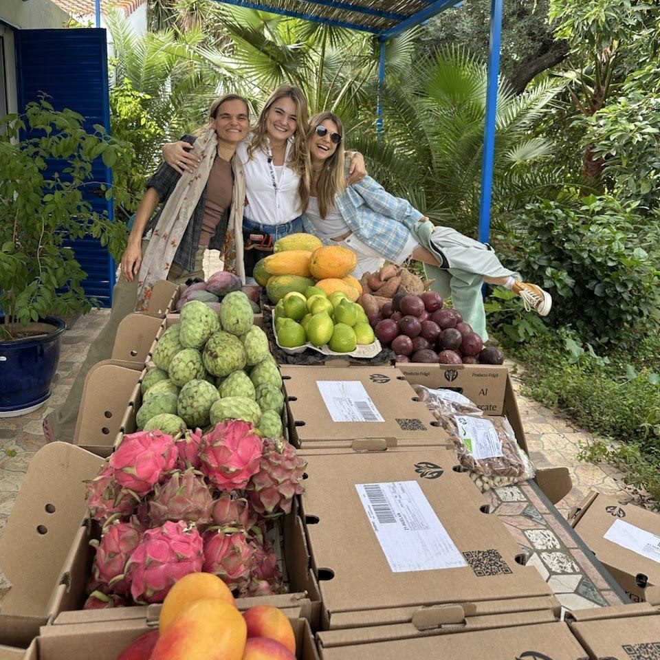 Trois femmes se serrent dans les bras et sourient derrière une table remplie de pitayas, de chérimoles, de papayes, de limes et de fruits de la passion